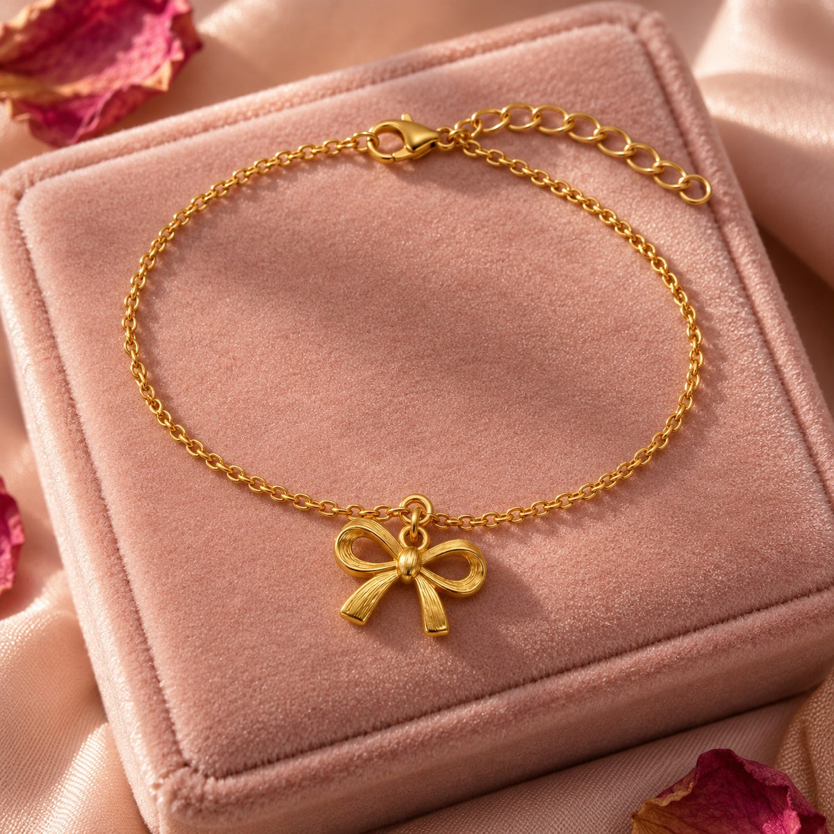 Gold bow charm bracelet styled on a blush velvet box with dried rose petals