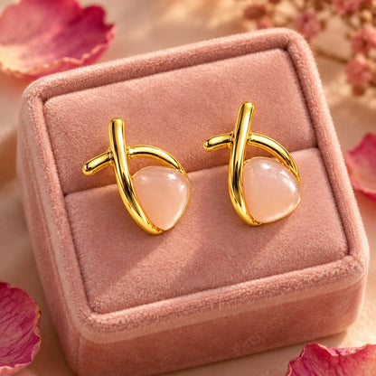 Gold crossover pink stone stud earrings on a blush velvet jewellery box with petals