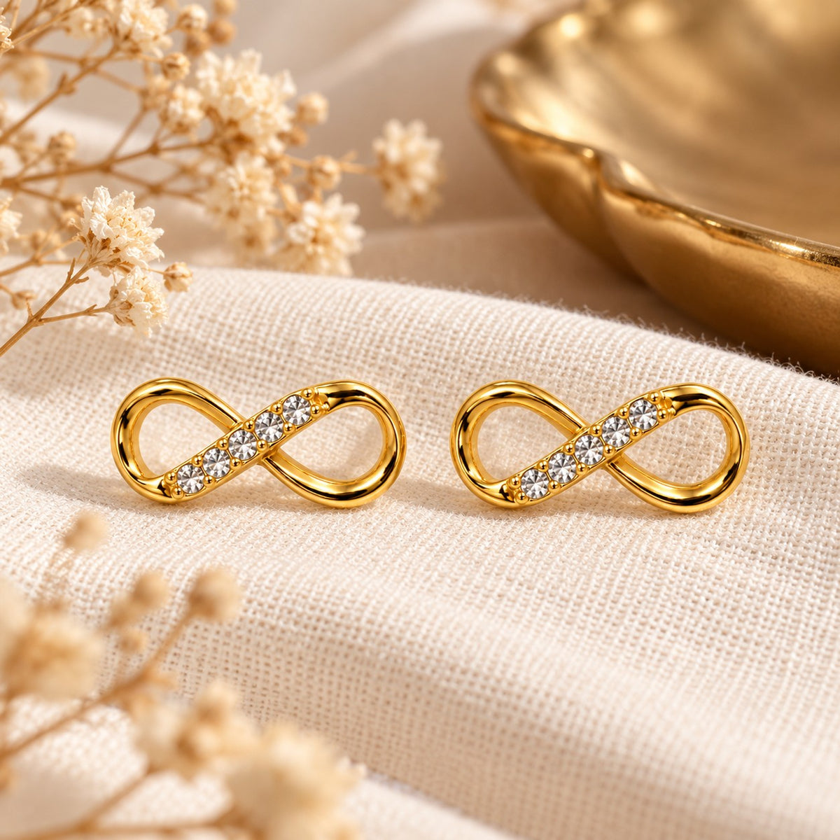 Gold infinity crystal stud earrings on cream linen with dried flowers