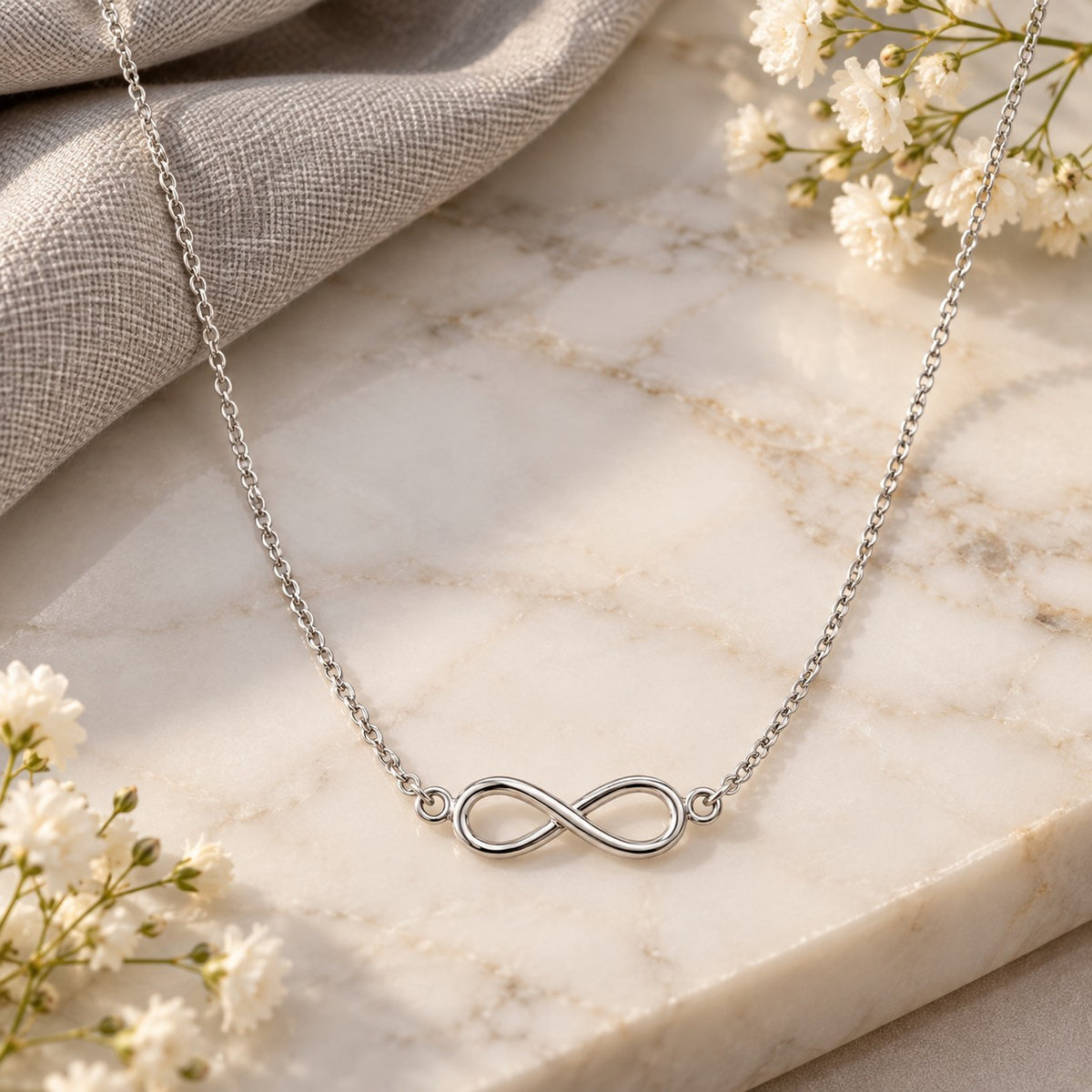 Silver infinity pendant chain necklace draped on white marble with soft flowers