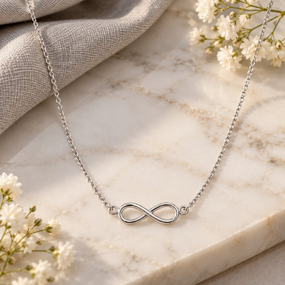 Silver infinity pendant chain necklace draped on white marble with soft flowers
